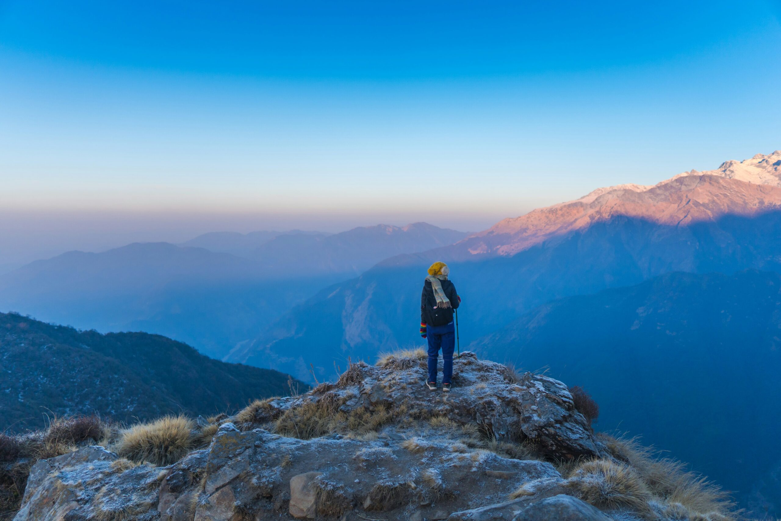 Person standing wearing winter hat at the top of mountain, looking into blue skies with many mountains.